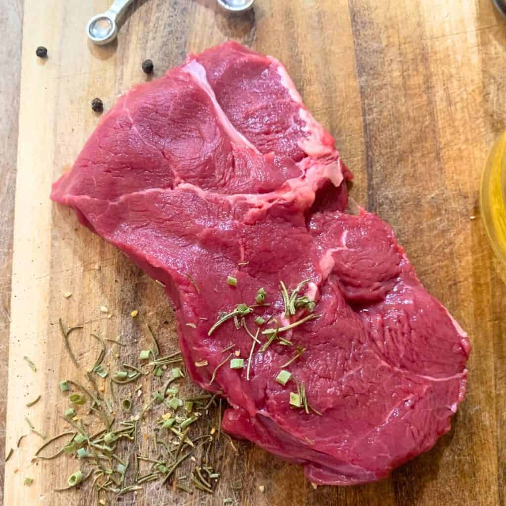 A raw, juicy steak on a wooden cutting board with salt and herbs on top.