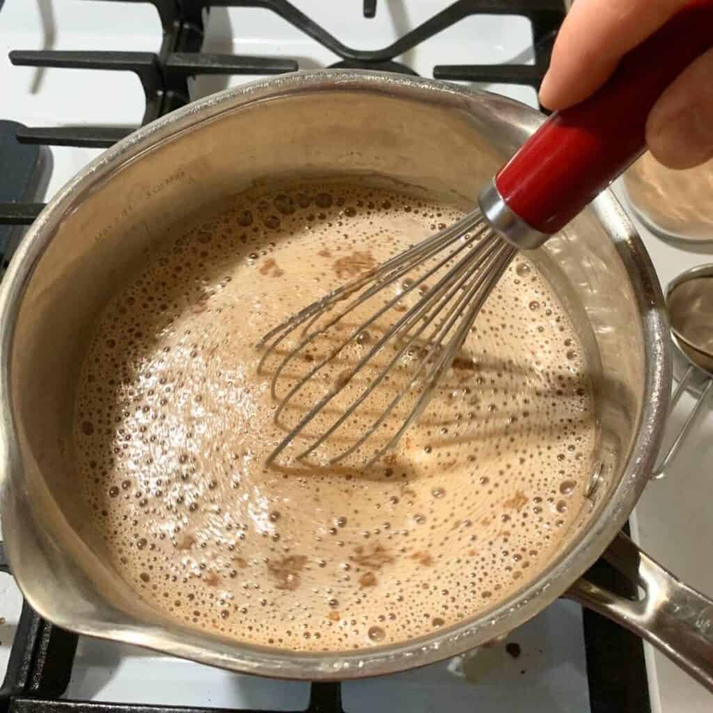 Woman stirring hot chocolate mixture in a medium pot on a stove, and using a wire whisk.