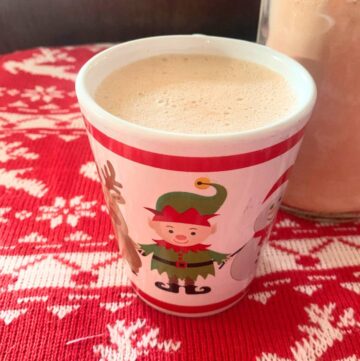 A white mug with a red stripe and elf sitting on a table covered with a red and white tablecloth. The mug has frothy hot chocolate inside. A glass jar of powdered cocoa mix sits behind the mug.