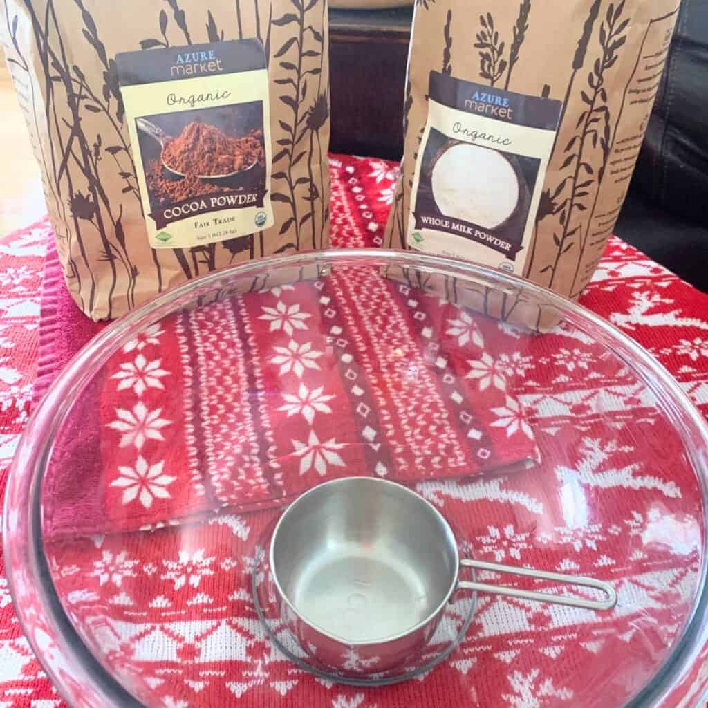 A clear glass bowl, stainless steel measuring cup, bag of organic cocoa powder, and bag of organic whole milk powder sitting on a table covered with a red and white table cloth.