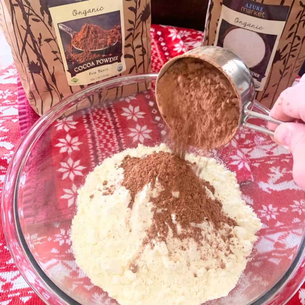 Woman adding cocoa powder and whole milk powder to a glass bowl.