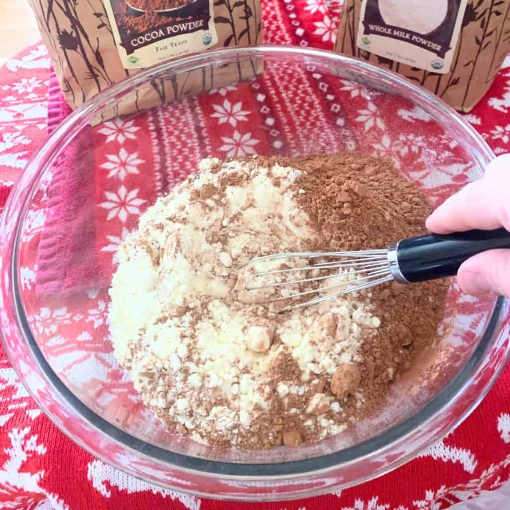 Woman using a wire whisk to mix cocoa powder and milk powder in a glass bowl.