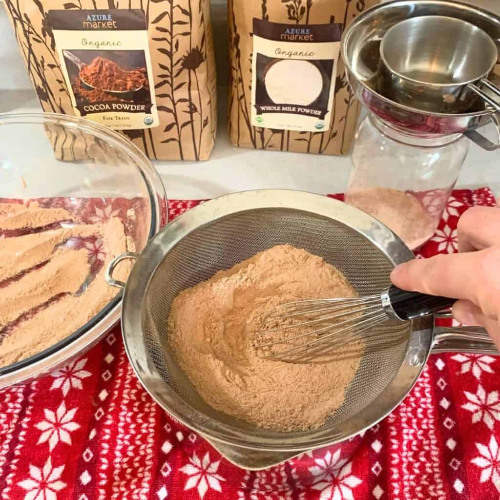 A woman stirring hot chocolate mix in a fine mesh sieve with a wire whisk.