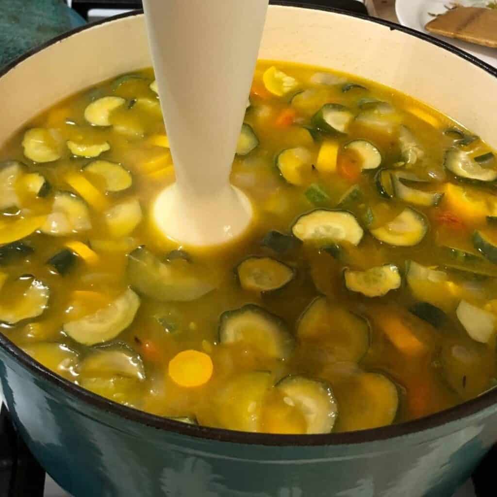 A turquoise Dutch oven on a stove with diced vegetables in chicken bone broth. A woman is using an immersion blender to puree the vegetables inside.