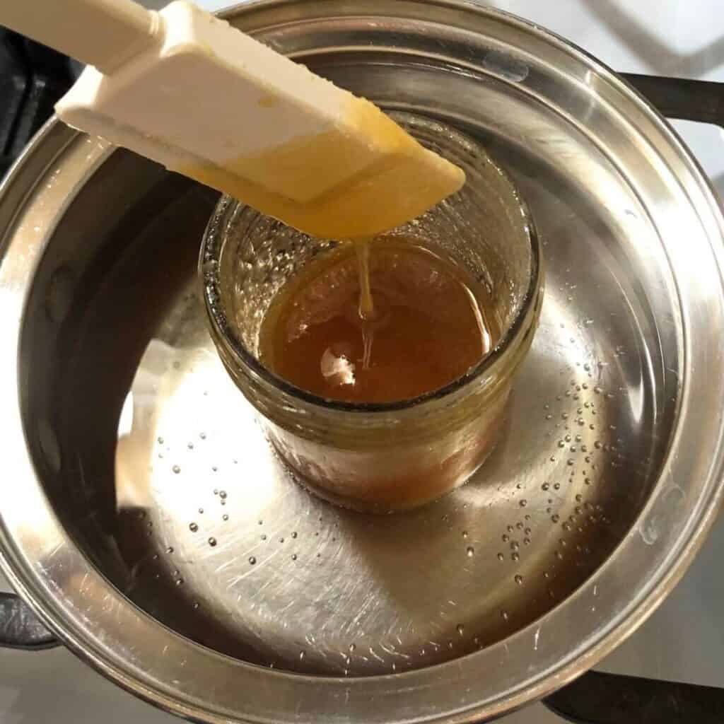 Woman stirring honey in a glass jar. The jar is in a small pot with some water, sitting on a stove.