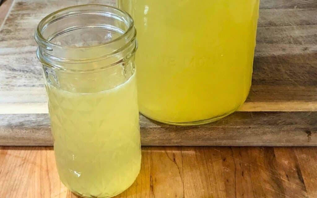 A glass of healthy honey lemonade sitting on a wooden cutting board in front of a half-gallon mason jar of lemonade.