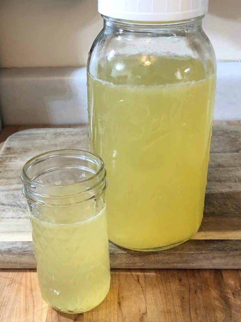 A glass of healthy honey lemonade sitting on a wooden cutting board in front of a half-gallon mason jar of lemonade.