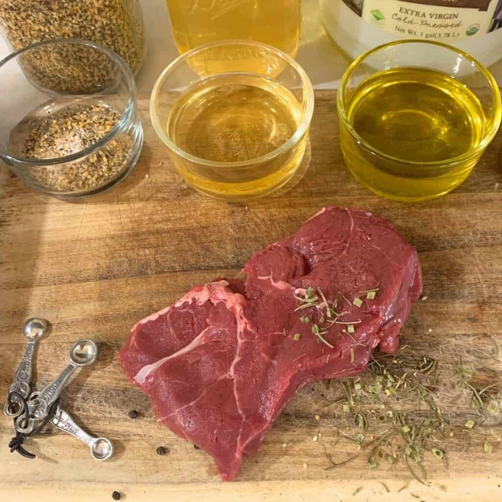 A beef steak on a wooden cutting board next to three ingredients of oil, vinegar, and Montreal steak seasoning.