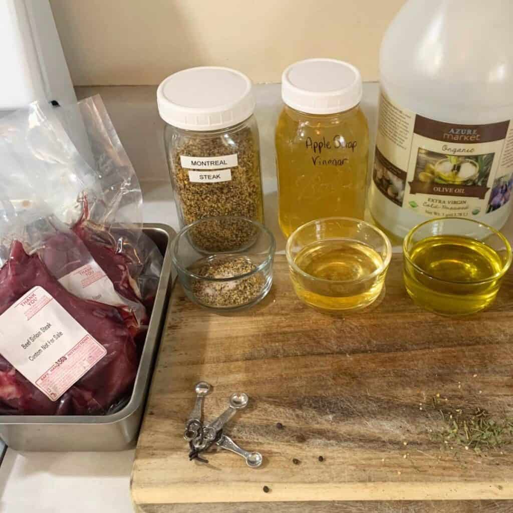 Ingredients for homemade steak marinade next to a wooden cutting board on a kitchen counter. Ingredients include oil, vinegar, Montreal steak seasoning, plus the beef steaks.