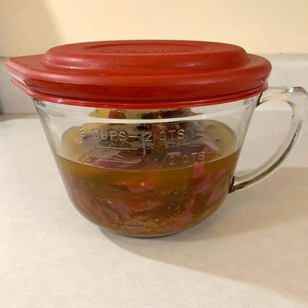 Steaks submerged in a large glass measuring cup of homemade steak marinade. It is covered with a red lid.