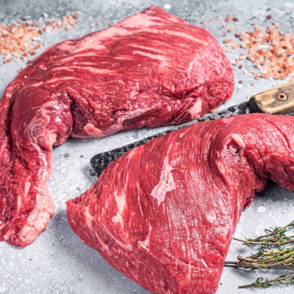 Two uncooked tri tip roasts on a counter with spices.