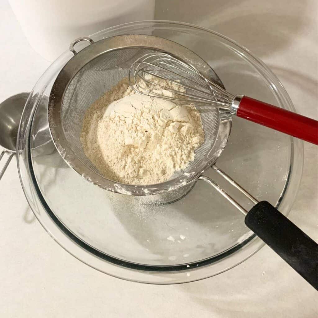 Freshly milled flour inside a fine mesh sieve set over a glass bowl. A balloon whisk is inside the sieve.