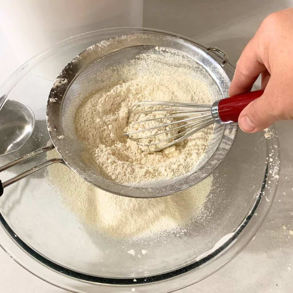 Woman stirring freshly milled flour inside a fine mesh sieve set over a glass bowl using a balloon whisk.