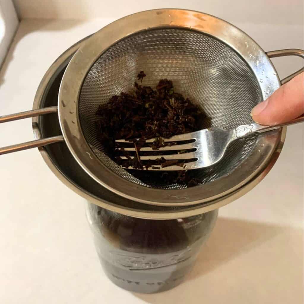 Woman squeezing from blackberry tea leaves in a fine mesh sieve using a fork.