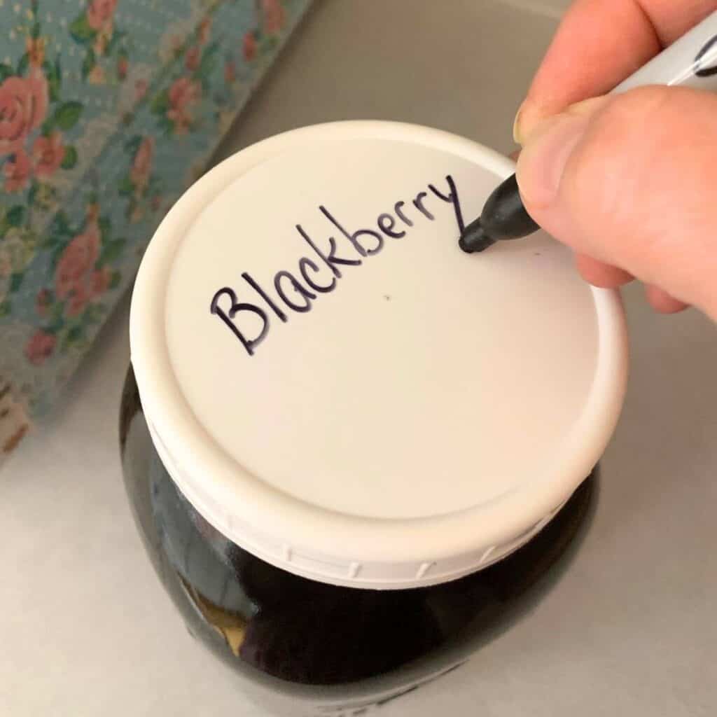 Woman writing "Blackberry tea" on the lid of a glass mason jar.