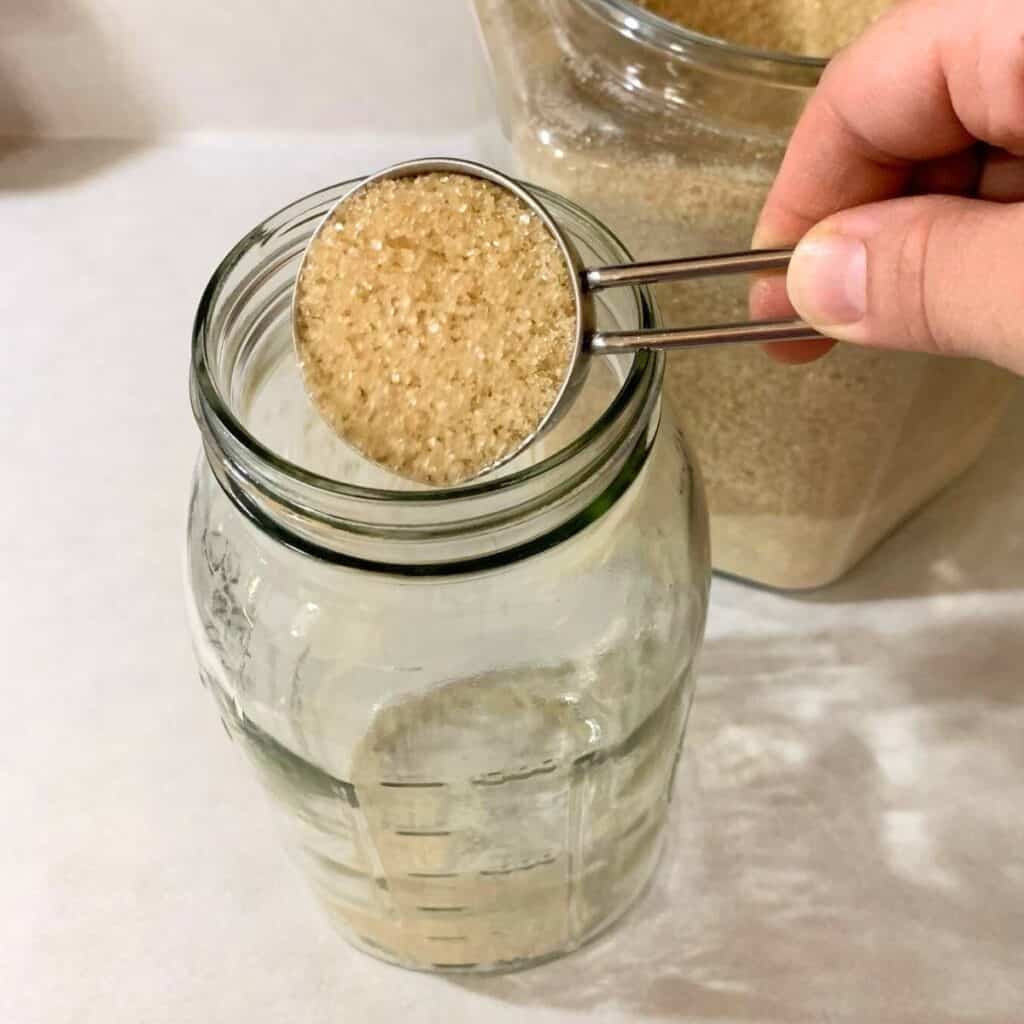 Woman adding a quarter cup of sugar to a half gallon glass mason jar.