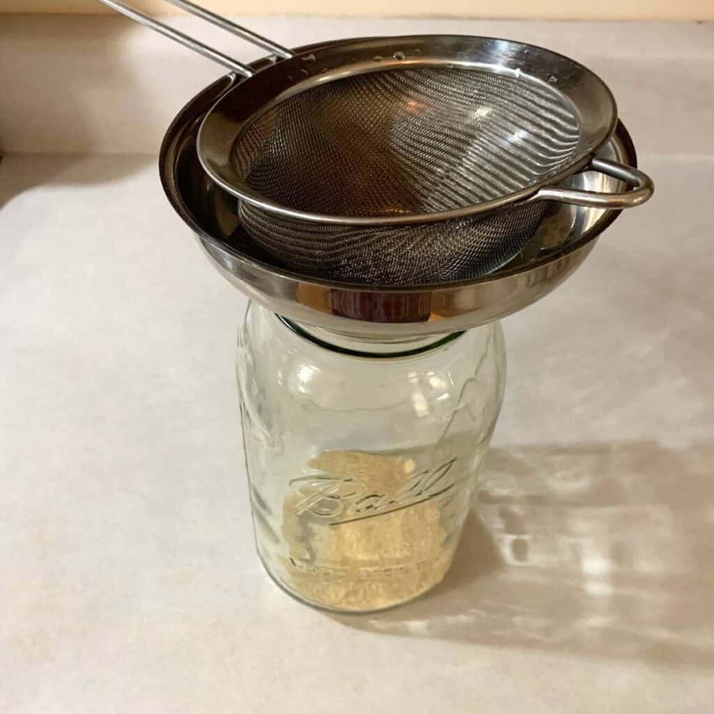 A half gallon glass mason jar with sugar in the bottom, with a stainless steel funnel and fine mesh sieve on top.