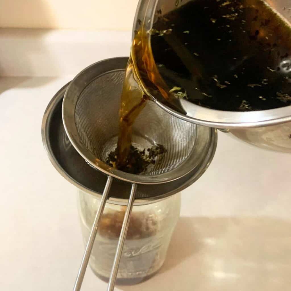 Woman pouring loose leaf blackberry tea through a fine mesh sieve and funnel into a glass mason jar.