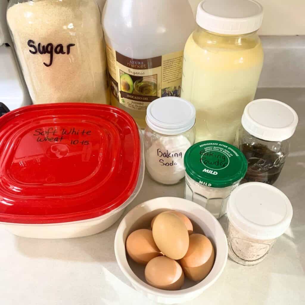 Ingredients for making the best homemade pancakes using freshly milled flour. On a counter are eggs, flour, sugar, avocado oil, milk, baking powder, baking soda, salt, and vanilla extract.