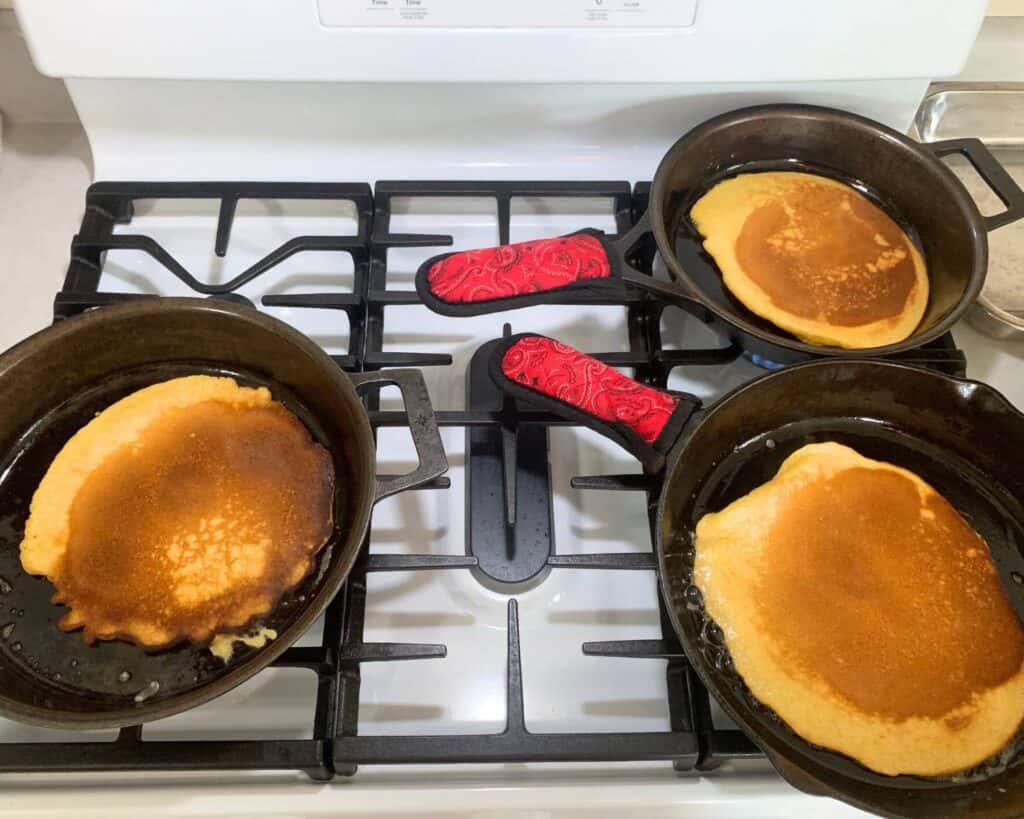 Three cast iron skillets on a gas stove with pancakes cooking in them.