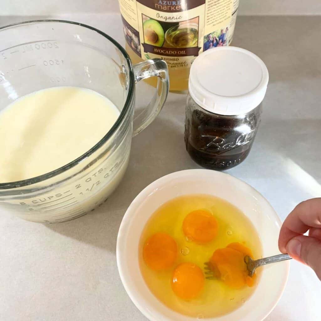 Woman scrambling eggs in a bowl. A mixing bowl of milk is behind it along with avocado oil and vanilla extract.