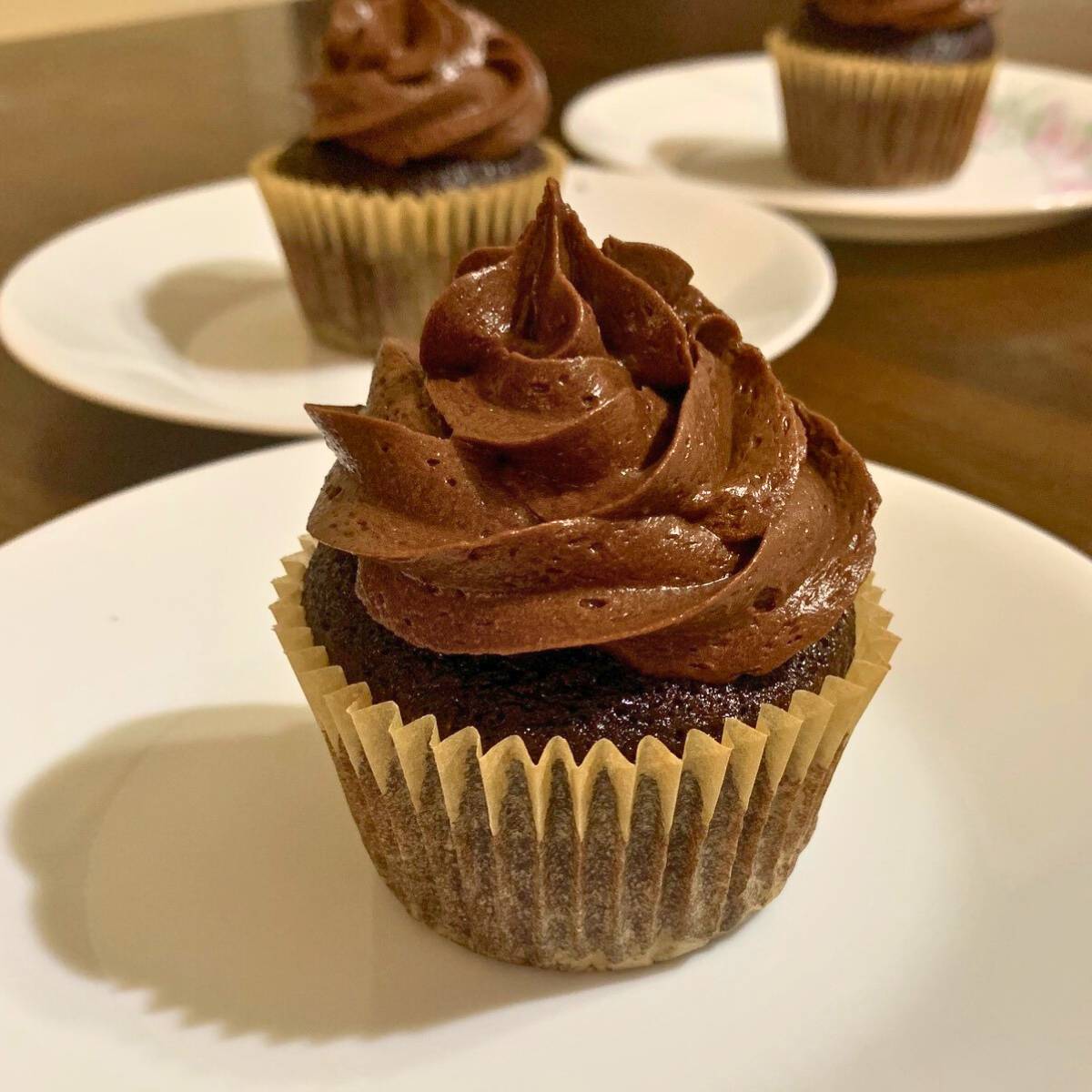 A chocolate cupcake with chocolate icing on a white plate sitting on a wood table.