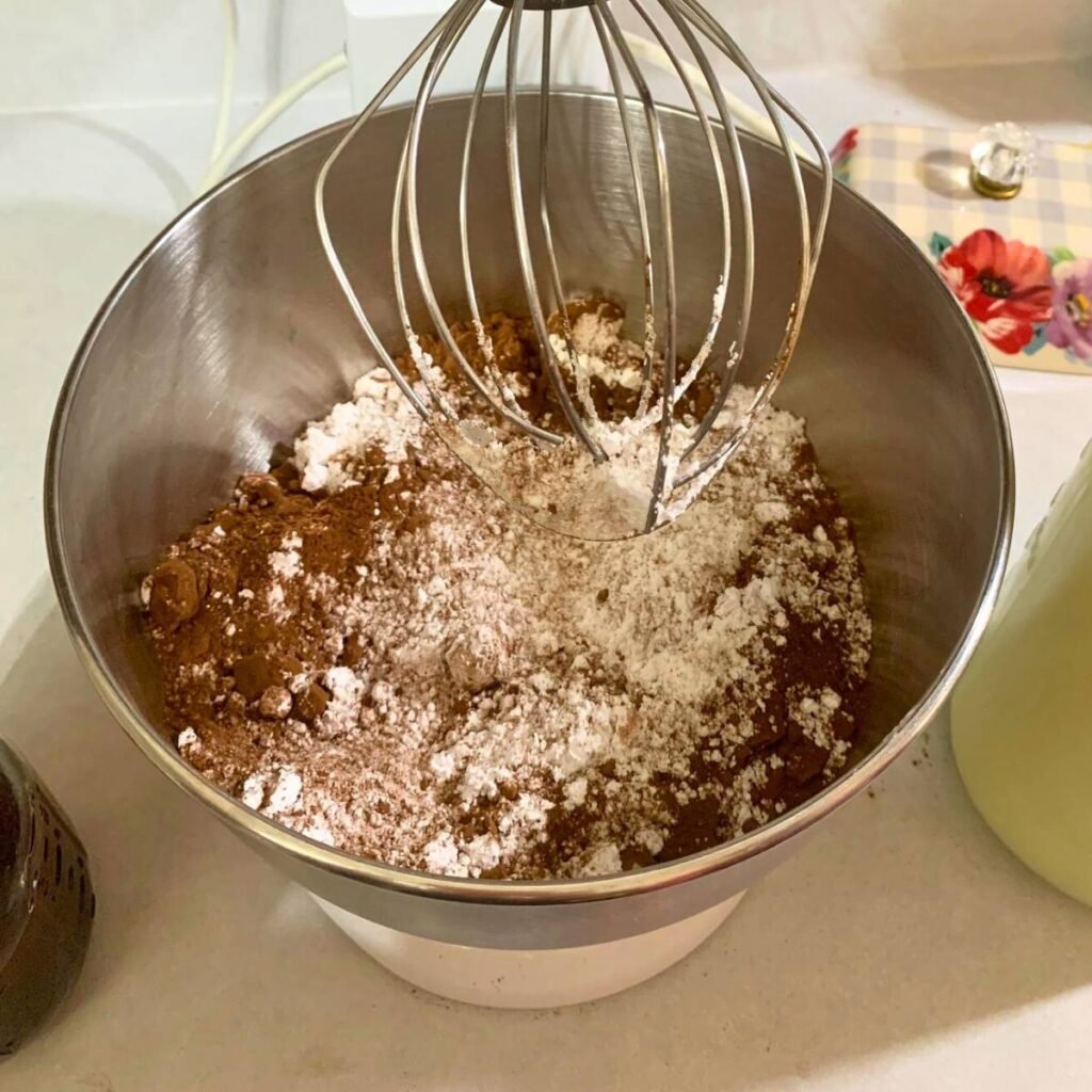 A stand mixer with a whisk attachment, with powdered sugar and cocoa powder inside the bowl.