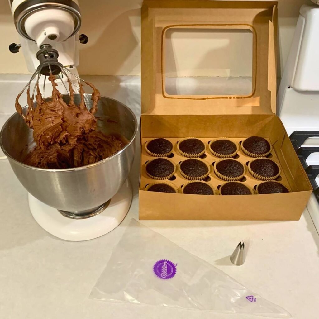 A stand mixer with chocolate icing inside sitting next to a brown box of chocolate cupcakes.