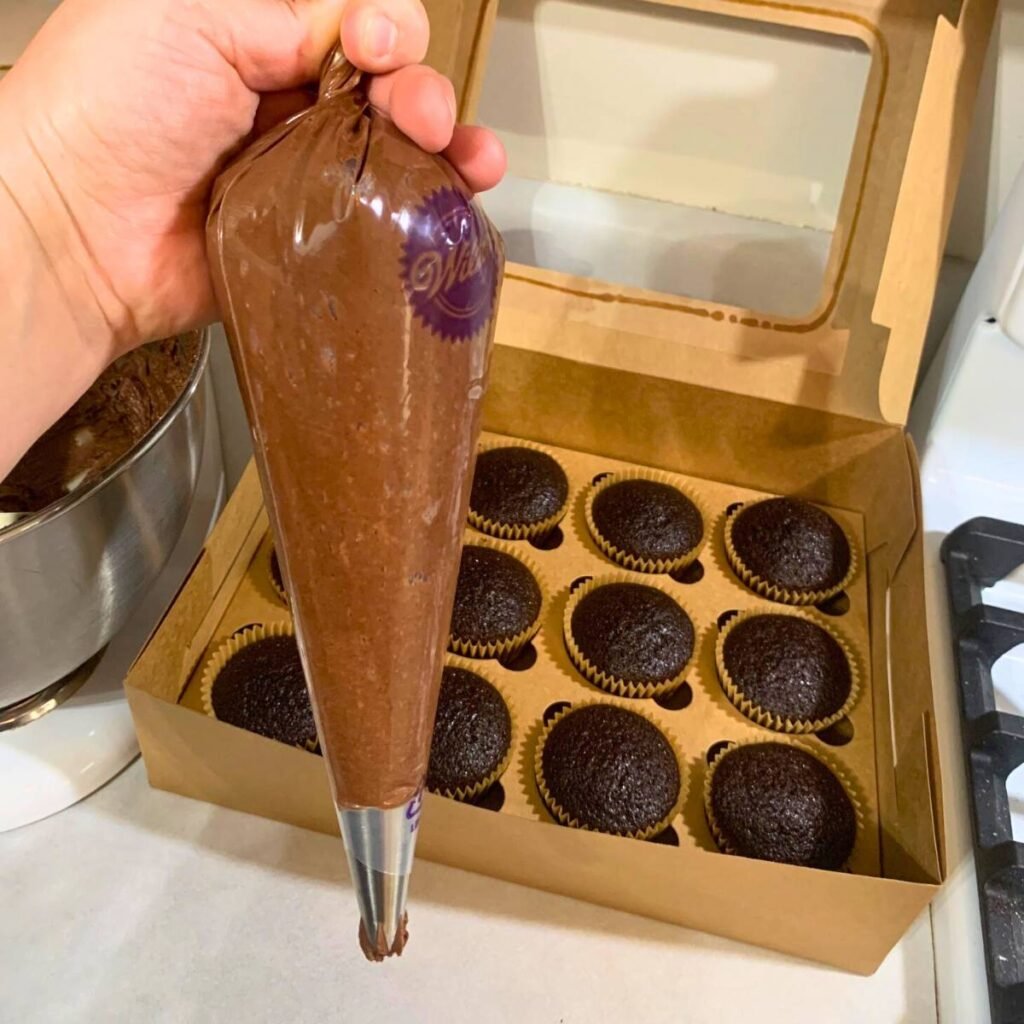 Woman holding a piping bag full of chocolate icing next to a dozen chocolate cupcakes inside a cupcake box.