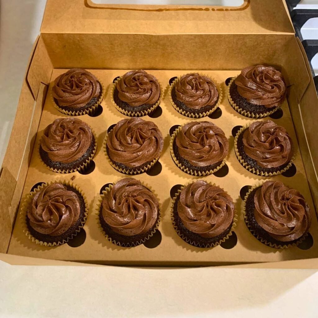 A dozen chocolate cupcakes with chocolate icing inside a disposable brown cupcake box.