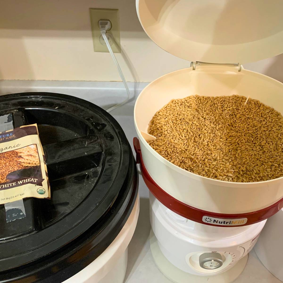A white Nutrimill grain mill with wheat berries inside, sitting on a kitchen counter.
