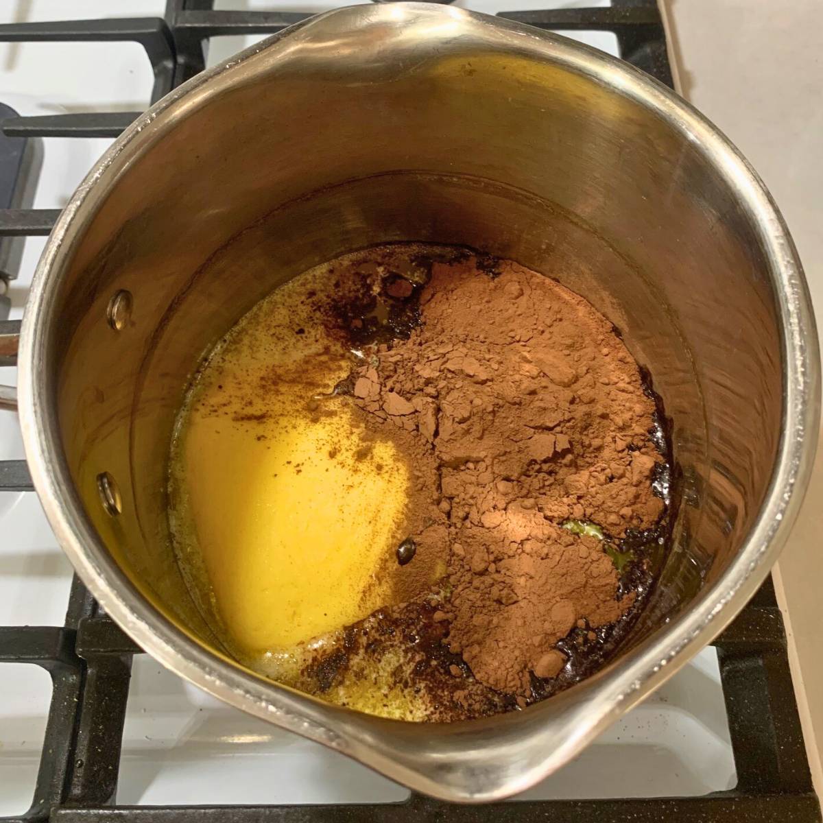 A stainless steel pot on a gas stove with melted butter and cocoa powder inside.