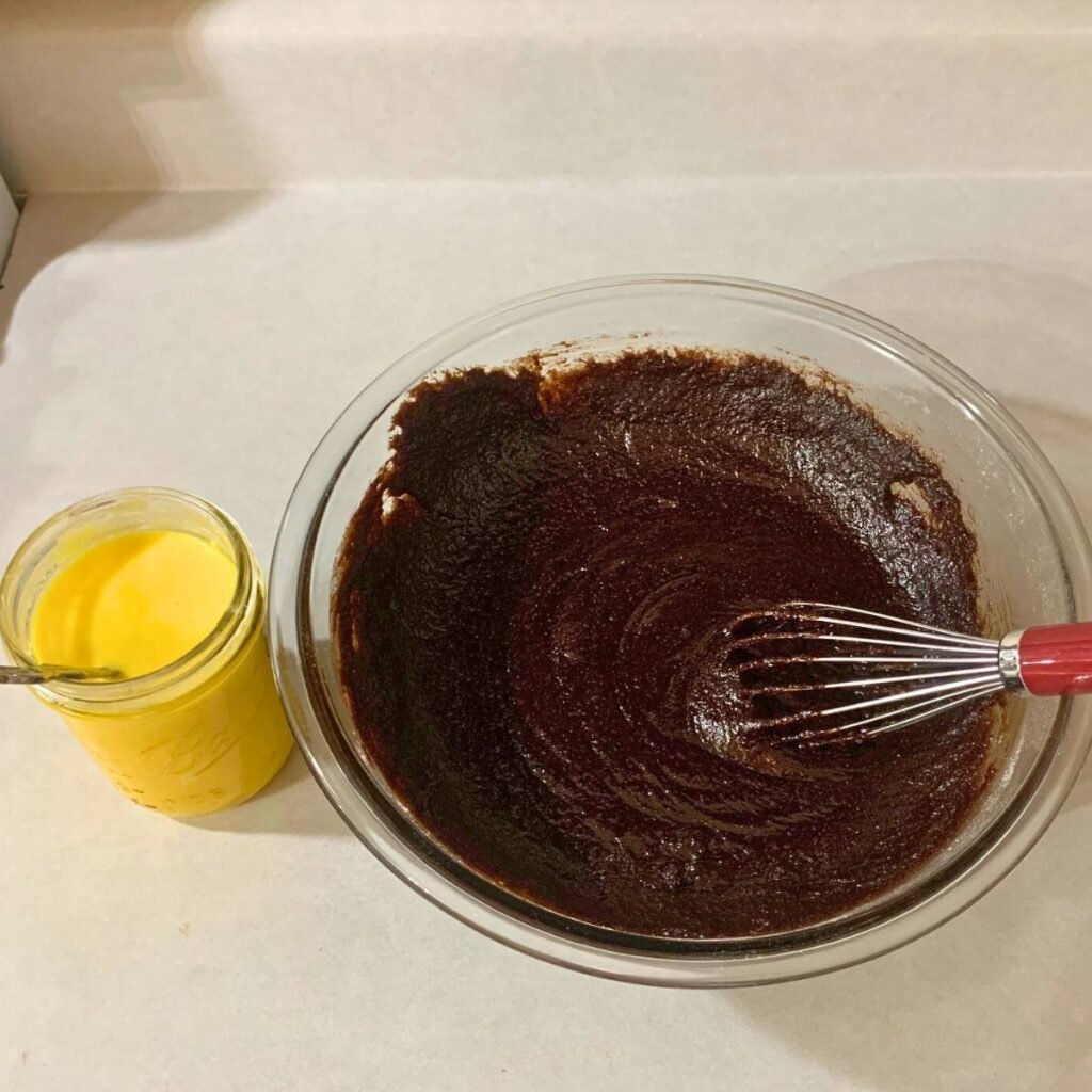 Woman whisking a chocolate cupcake batter in a glass bowl with a cup of egg mixture sitting next to it on a counter.