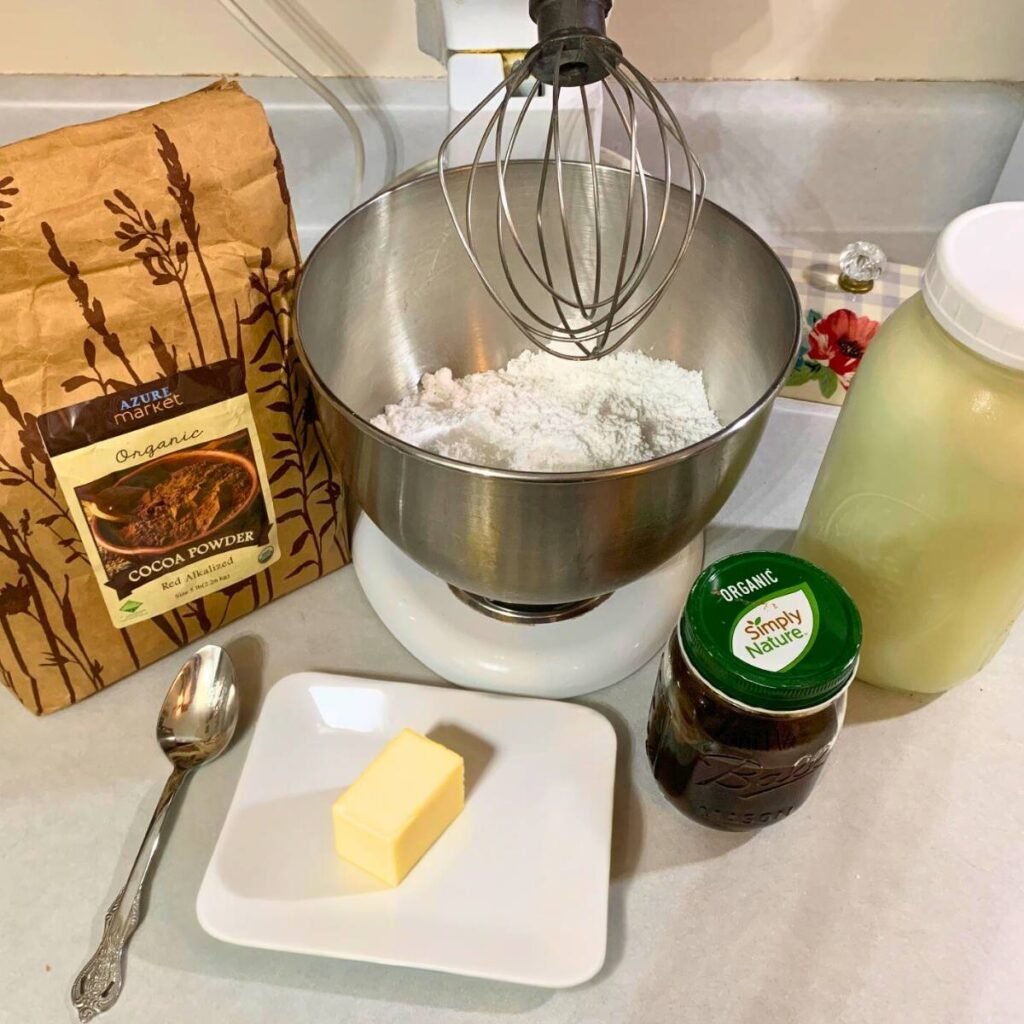 Ingredients for making chocolate icing including a stand mixer with powdered sugar in the bowl, a bag of cocoa powder, a half stick of butter, vanilla extract, and milk sitting on a counter.