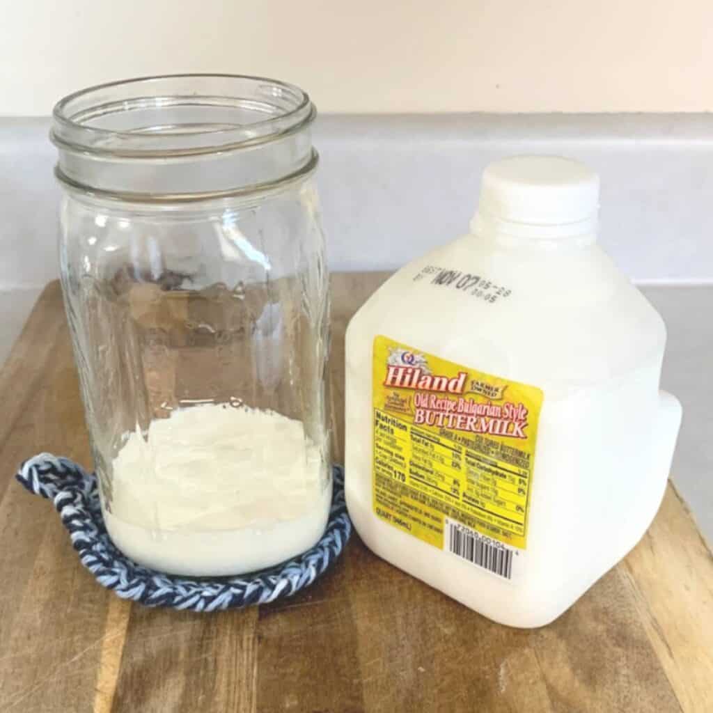 A quart mason jar with a half cup of buttermilk inside sitting next to a jug of buttermilk on a wood cutting board.