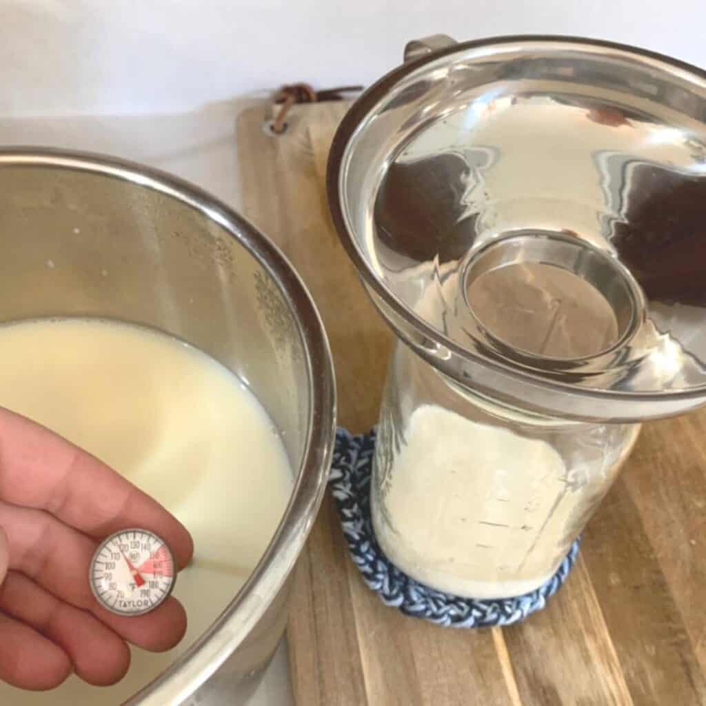 Woman holding a thermometer in a pot of hot milk and a quart mason jar with a stainless steel funnel sitting on the rim of the jar.