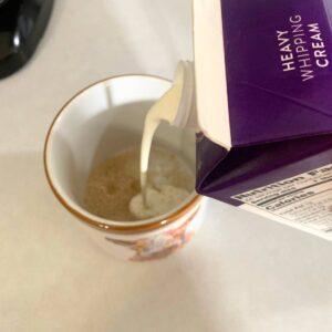 Woman adding heavy cream to a coffee cup with sugar and vanilla extract inside.