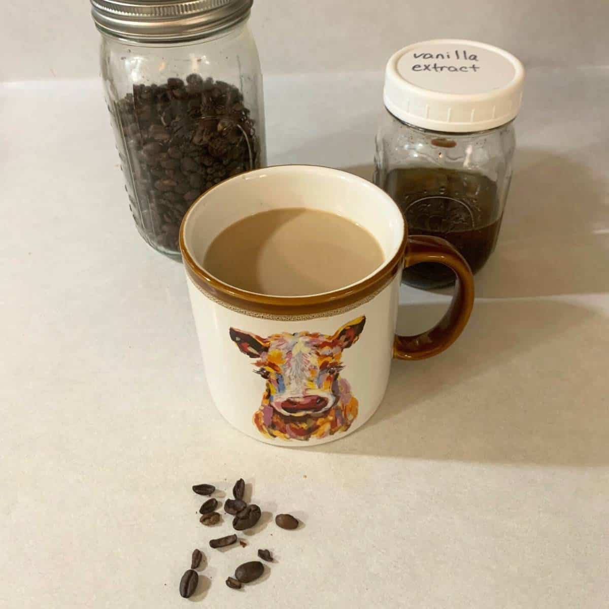 A mug of coffee with a picture of a cow on it. The coffee is in front of a mason jar of coffee beans and homemade vanilla extract. A few coffee beans are on the counter.