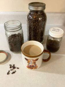 Glass mason jars of whole roasted coffee beans and homemade vanilla extract behind a mug of coffee with a picture of a cow on it.