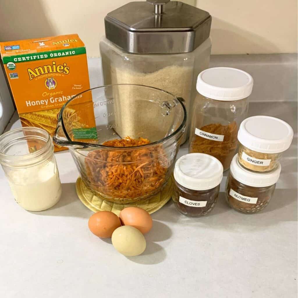 Ingredients sitting on a counter including graham crackers, sugar, shredded carrots, eggs, milk, cinnamon, ginger, nutmeg, and cloves.