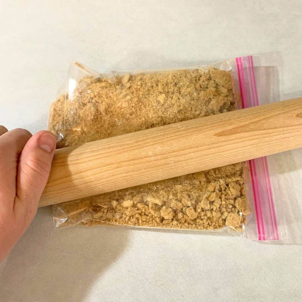 Woman crushing graham crackers in a zip top bag with a rolling pin.