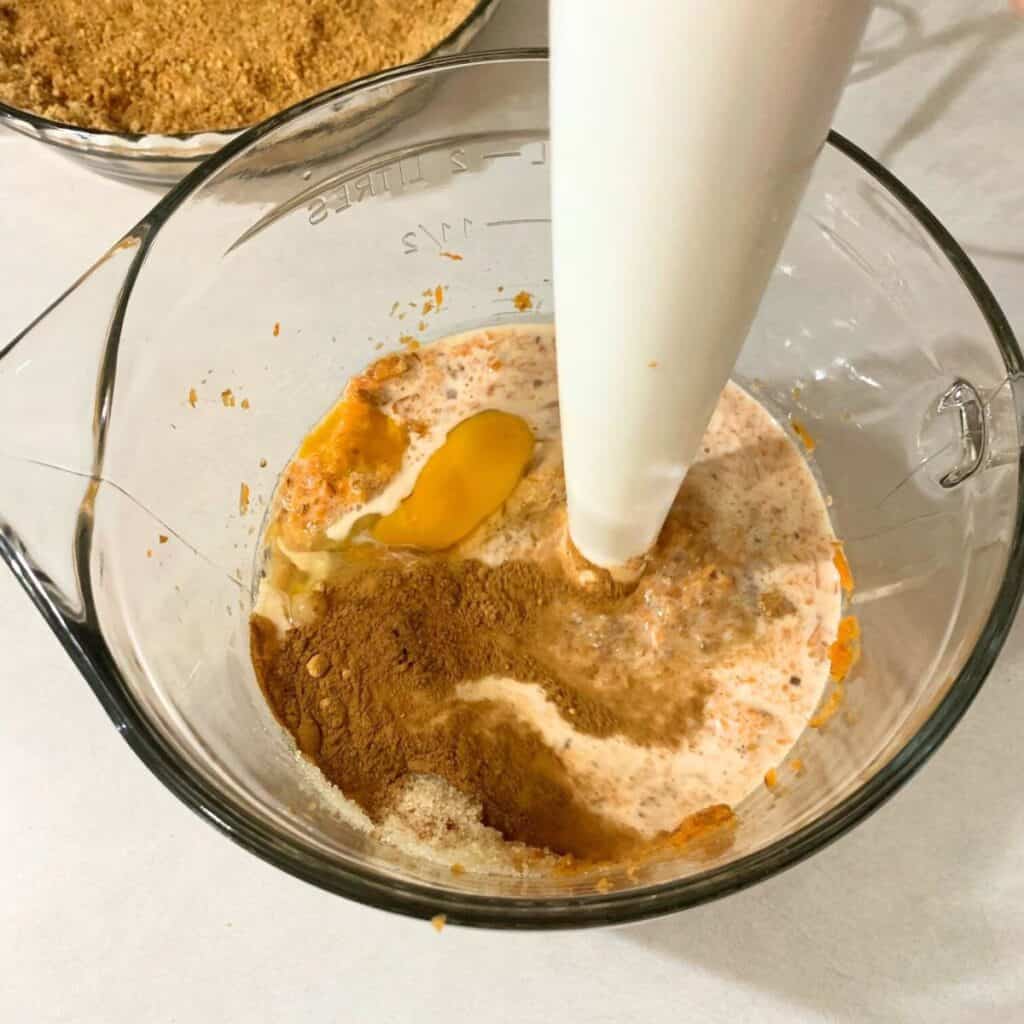 Woman mixing together steamed carrots, milk, sugar, eggs, cinnamon, ginger, nutmeg, and cloves.