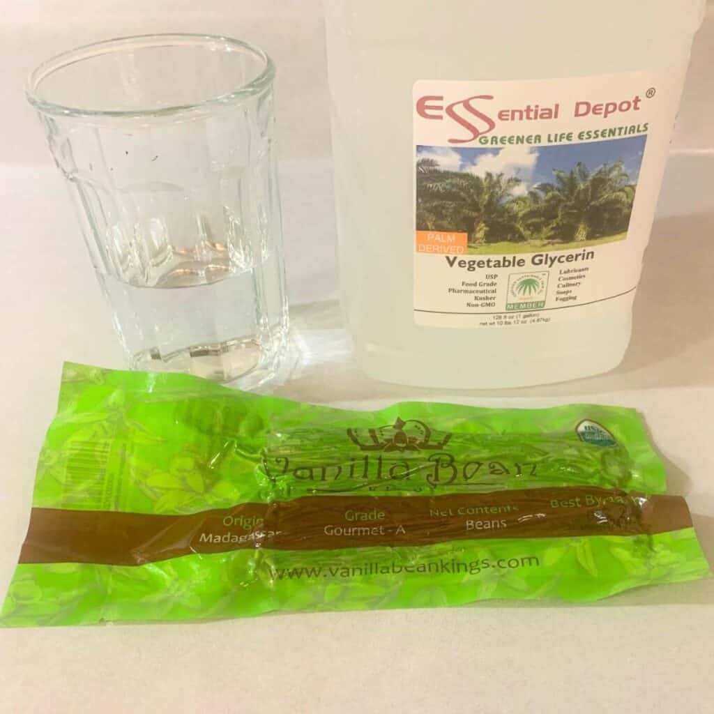 A glass of filtered water, a jug of food grade vegetable glycerin, and a package of Madagascar vanilla beans sitting on a kitchen counter.