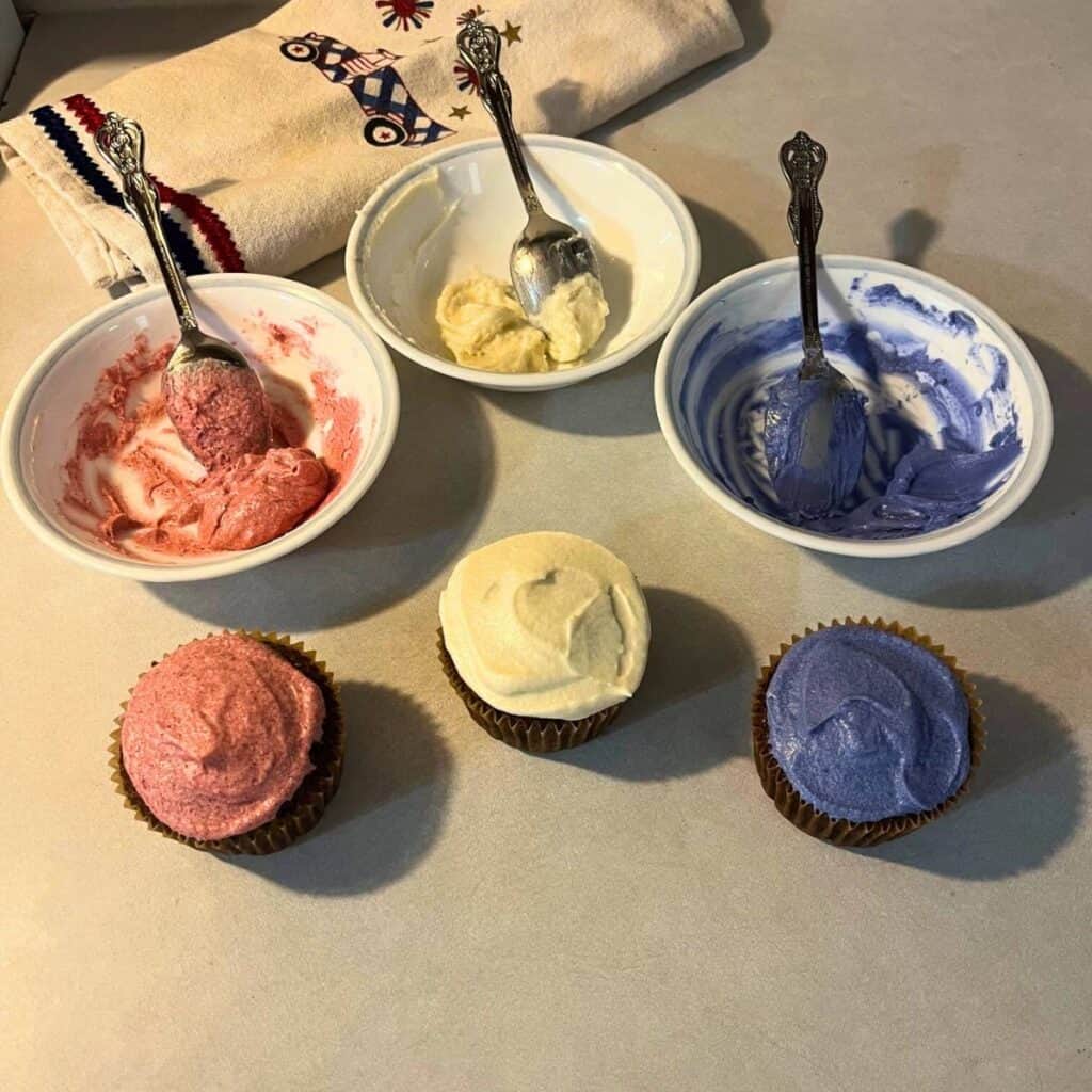 Three chocolate cupcakes sitting on a kitchen counter with red, white, and blue icing along with a white kitchen towel with Fourth of July decorations.