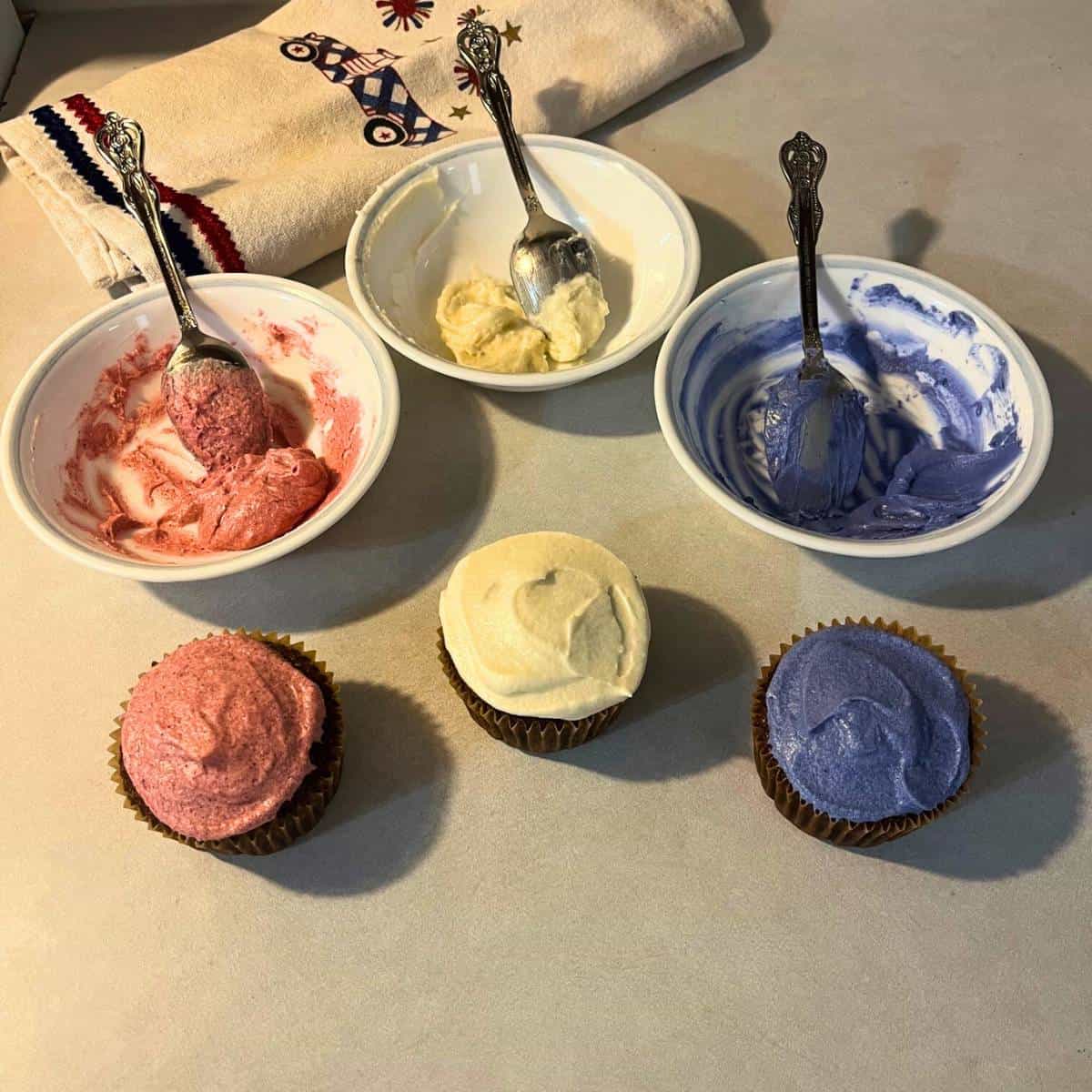 Three chocolate cupcakes sitting on a kitchen counter with red, white, and blue icing along with a white kitchen towel with Fourth of July decorations.