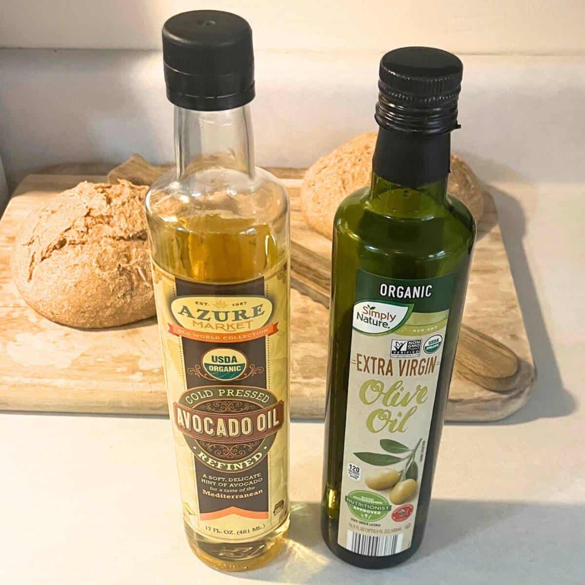 Jars of organic olive oil and organic avocado oil on a counter in front of two loaves of artisan bread on a wood cutting board.