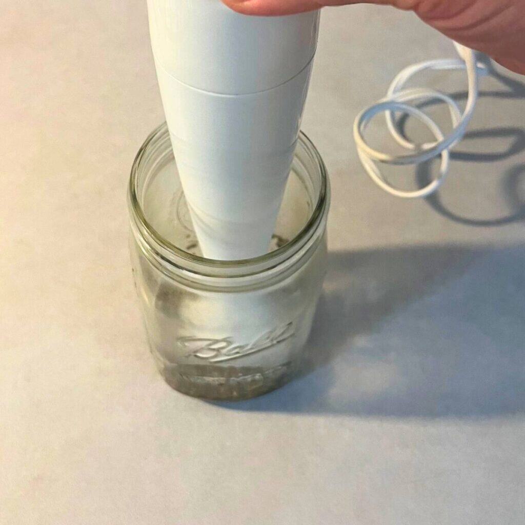 Woman using a white immersion blender to blend together simmering water and dried comfrey leaves in a glass mason jar.