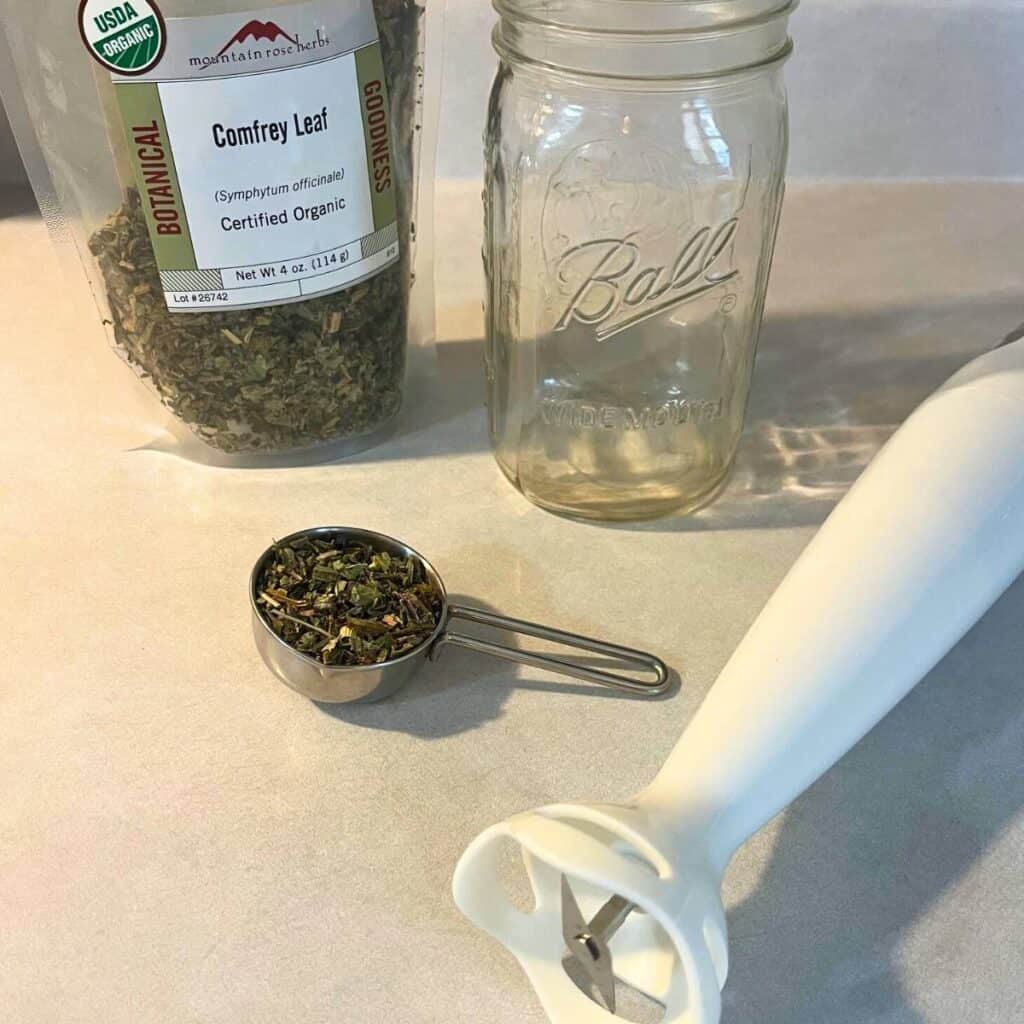 Items needed to create a comfrey poultice sitting on a counter. Included are dried organic comfrey leaves, a quart jar, and an immersion blender.