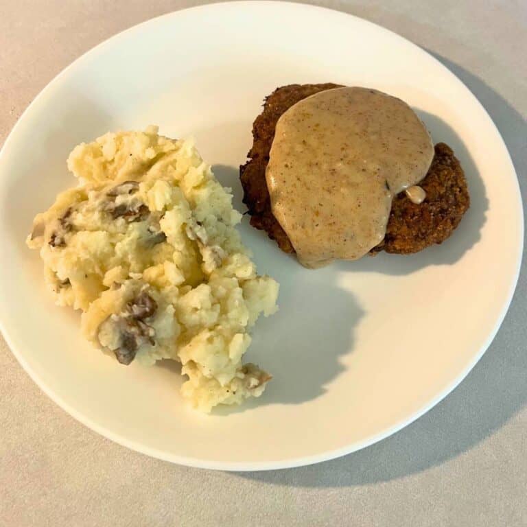 A crispy country fried steak patty with gravy on top next to mashed potatoes on a white plate.