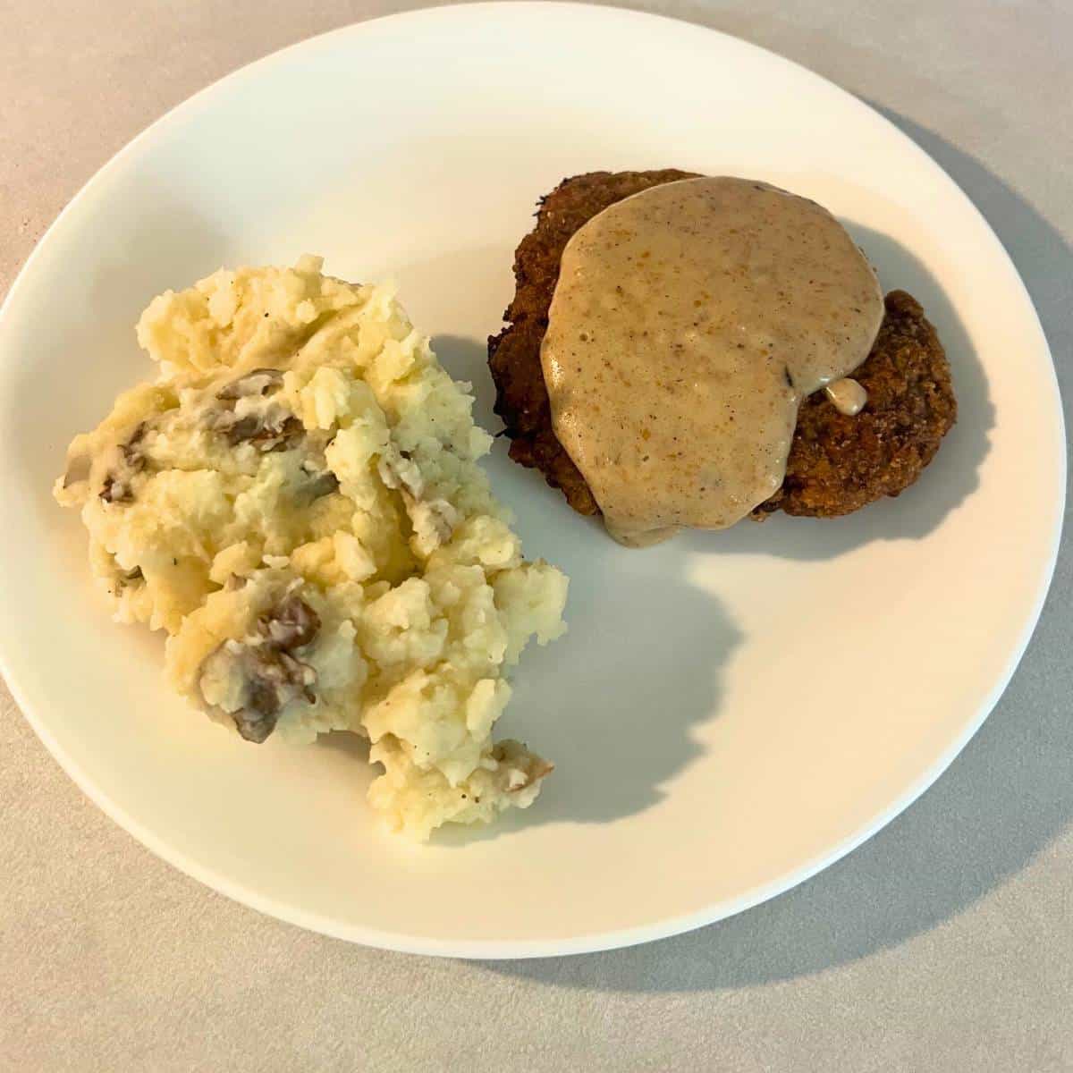 A crispy country fried steak patty with gravy on top next to mashed potatoes on a white plate.